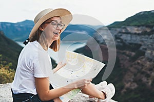 Laughing young blonde tourist holds in hands map of  landscape and looks views top of mountain landscape, cute smiling girl in hat