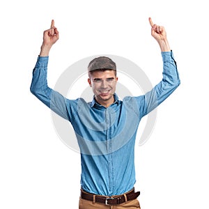 Laughing young casual man pointing fingers up above his head
