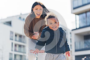 Laughing young caucasian mother chasing her little giggly biracial toddler son. Sunny day in the city during the weekend