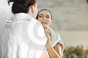 Laughing young girl applying lipstick on lips looking at her reflection in wall mirror at home