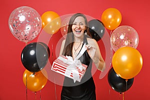 Laughing young girl in black dress celebrate, opening red box with gift present on bright red background air balloons