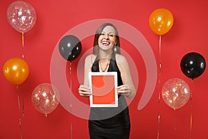 Laughing young girl in black dress celebrating, holding tablet pc computer with blank black empty screen on bright red