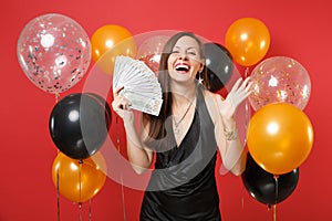 Laughing young girl in black dress spreading hands, holding bundle lots of dollars, cash money on red background air