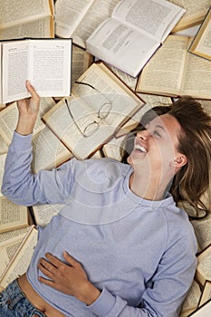 A laughing young girl in a blue sweater and jeans lies on a pile of open books reading. Education and knowledge. Vertical