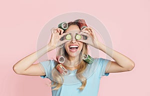 Laughing young girl in curlers puts pieces of cucumber on eyes