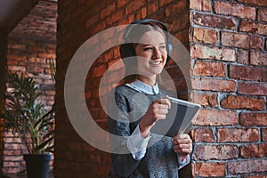 Laughing young girl dressed in an elegant gray dress leaning against brick wall, listening music in headphones and