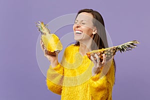 Laughing young girl in fur sweater keeping eyes closed hold halfs of fresh ripe pineapple fruit isolated on violet