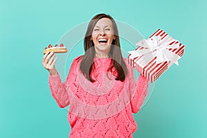 Laughing young girl in knitted pink sweater hold eclair cake red striped present box with gift ribbon isolated on blue