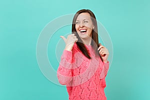 Laughing young girl in knitted pink sweater pointing thumb behind her back isolated on blue turquoise wall background