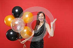 Laughing young girl in little black dress celebrating spreading hands holding air balloons isolated on red background