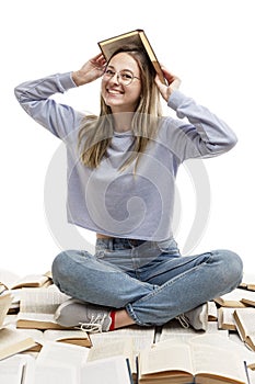 A laughing young girl sits on a pile of opened books and holds a book on her head. Education and knowledge. Isolated on a white