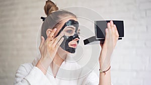 Laughing young girl with a towel on her head puts a black mask on her face, day spa