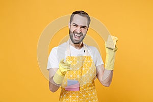 Laughing young man househusband in apron rubber gloves hold cleaning sponge rag while doing housework isolated on yellow