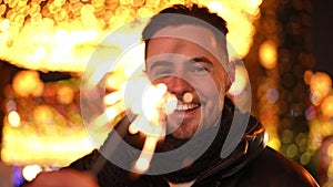 Laughing young man with sparklers looking at camera smiling talking standing in urban city at night. Portrait of happy