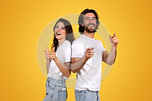 Laughing young woman and bearded man standing back to back, playfully pointing fingers at the camera