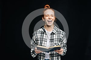 Laughing young woman with closed eyes college student holding opened book on black isolated background.