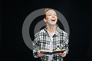 Laughing young woman college student with closed eyes holding opened book on black background.