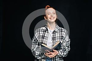 Laughing young woman college student holding opened book and looking at camera on black background