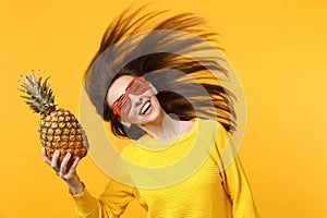 Laughing young woman in funny glasses with flowing hair holding fresh ripe pineapple fruit isolated on yellow orange