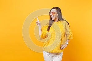 Laughing young woman in fur sweater, heart orange glasses pointing index finger aside on copy space isolated on bright