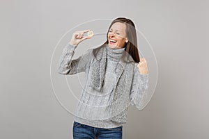 Laughing young woman in gray sweater, scarf clenching fist like winner hold bitcoin, future currency isolated on grey