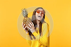 Laughing young woman in heart glasses holding in hands fresh ripe pineapple fruit isolated on yellow orange background