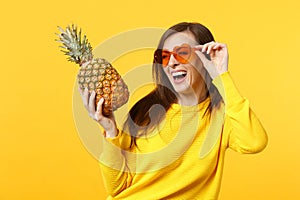 Laughing young woman in heart glasses looking aside hold fresh ripe pineapple fruit isolated on yellow orange background