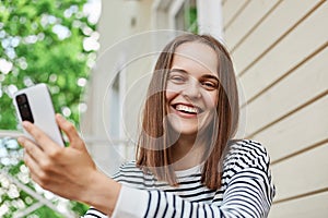 Laughing young woman holding her smartphone in hands while sitting on porch of house using device to send message or browsing