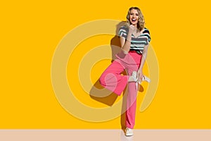 Laughing young woman in pink high waisted baggy pants and sneakers is posing on one leg in front of yellow wall
