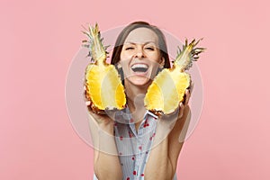 Laughing young woman in summer clothes holding halfs of fresh ripe pineapple fruit isolated on pink pastel wall