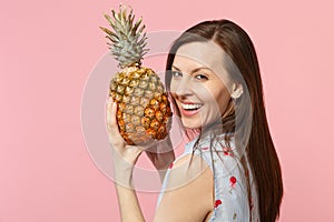 Laughing young woman in summer clothes holding in hands fresh ripe pineapple fruit isolated on pink pastel wall