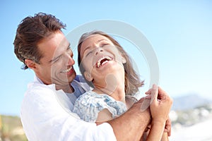 Laughter is the secret to love. A mature man embracing his happy wife from behind as they stand on the beach.