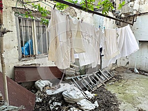 Laundry hangs on line in dilapidated city yard with overgrown leaves, broken furniture and trash, depicting neglect and