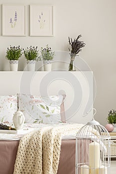 Lavender flowers on bedhead of bed with blanket in bright bedroom interior with candles and posters. Real photo