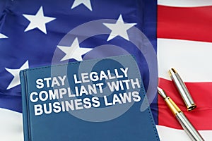On the US flag lies a pen and a book with the inscription - STAY LEGALLY COMPLIANT WITH BUSINESS LAWS