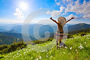 On the lawn in mountains landscapes the hipster girl in dress, stockings and straw hat stays watching the sky with clouds.