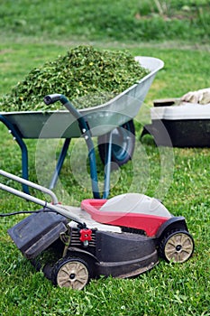 Lawn mower, grass container and a garden wheelbarrow full of trimmed grass on the mowed lawn