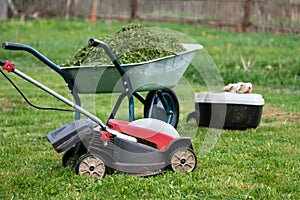 Lawn mower, grass container and a garden wheelbarrow full of trimmed grass on the mowed lawn