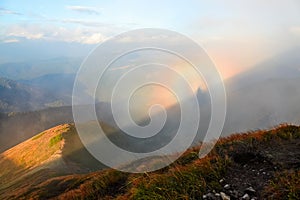 From the lawn with orange grass opens a panorama of high mountains, blue sky with clouds and a Brocken Spectre in the fog.