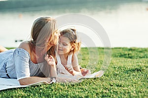 Lay down, the grass is magnificent. Photo of young mother and her daughter having good time on the green grass with lake