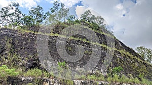 Layered rock face covered in sparse vegetation under a partly cloudy sky. The geological strata are visible, showing photo