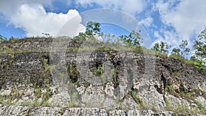 Layered rock face covered in sparse vegetation under a partly cloudy sky. The geological strata are visible, showing photo