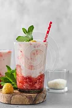 Layered strawberry and raspberry smoothie or milkshake in glasses decorated with mint, raspberry and coconut flakes on grey table