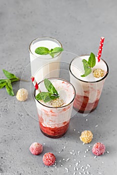 Layered strawberry and raspberry smoothie or milkshake in glasses decorated with mint, raspberry and coconut flakes on grey table