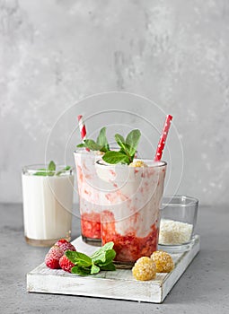 Layered strawberry and raspberry smoothie or milkshake in glasses decorated with mint, raspberry and coconut flakes on grey table