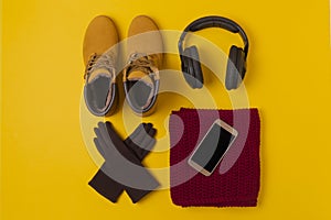 Layout of autumn clothes and accessories in warm colors on a yellow background. Gloves, headphones boots, phone and scarf flat lay