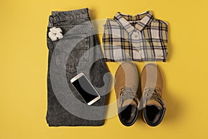 Layout of autumn clothes and accessories in warm colors on a yellow background. Jeans, shirt, boots and phone flat lay nearby