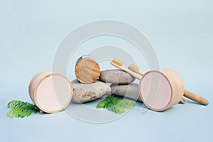 Layout of babmboo cans, cosmetic jar with wooden cover, two bamboo toothbrushes on grey pebbels with few green leaves and light