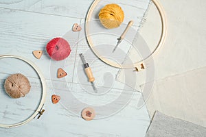 Layout of embriodery hoops, punch needles, balls of yarn, linnen fabric and wooden buttons on white wooden floor. Instruments and