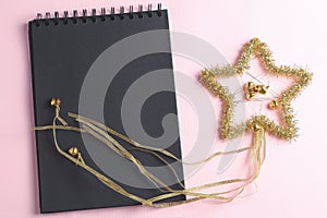 Layout for holiday to-do list. black notebook and star of tinsel on pink background. top view